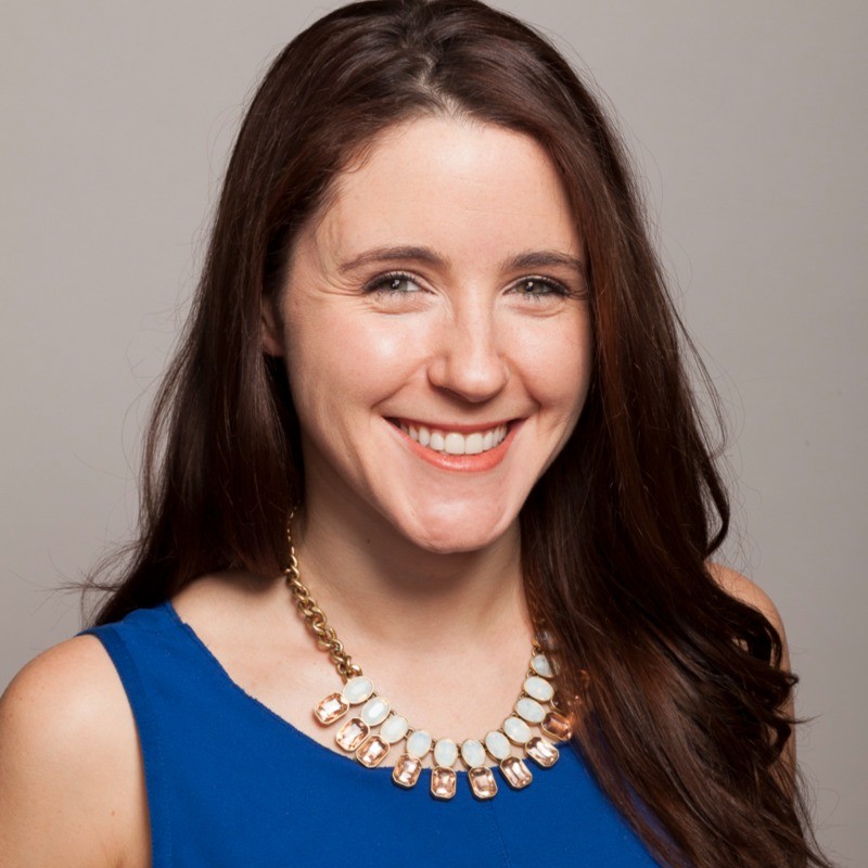 Smiling woman with chest-length dark brown hair, blue dress, and large necklace.